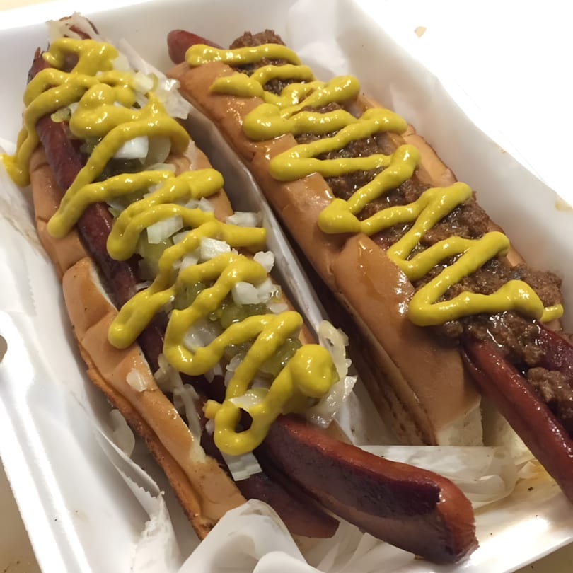 Two Jordan's Hot Dogs & Mac hot dogs in a white container, one with mustard and onions, the other with chili and mustard.
