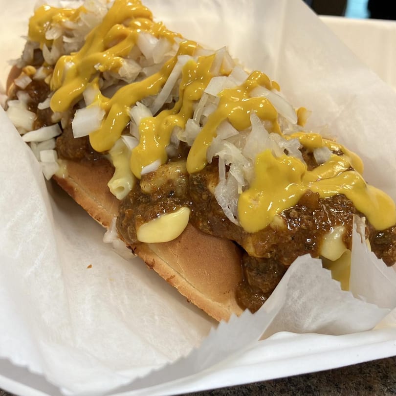 A Jordan's Hot Dogs & Mac chili dog with mustard, chopped onions, and macaroni in a paper tray.