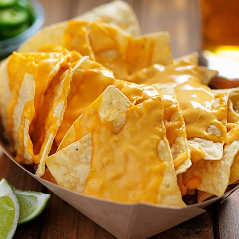 Cheesy nachos in a brown paper tray, with lime wedges and jalapeños in the background.